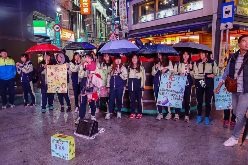 Taiwan-Taipei-Ximending-Ramen - These girls are protesting something with song. Their signs depict diamonds, hand bags, lipstick, and the King of Thailand? I cant read the traditiona